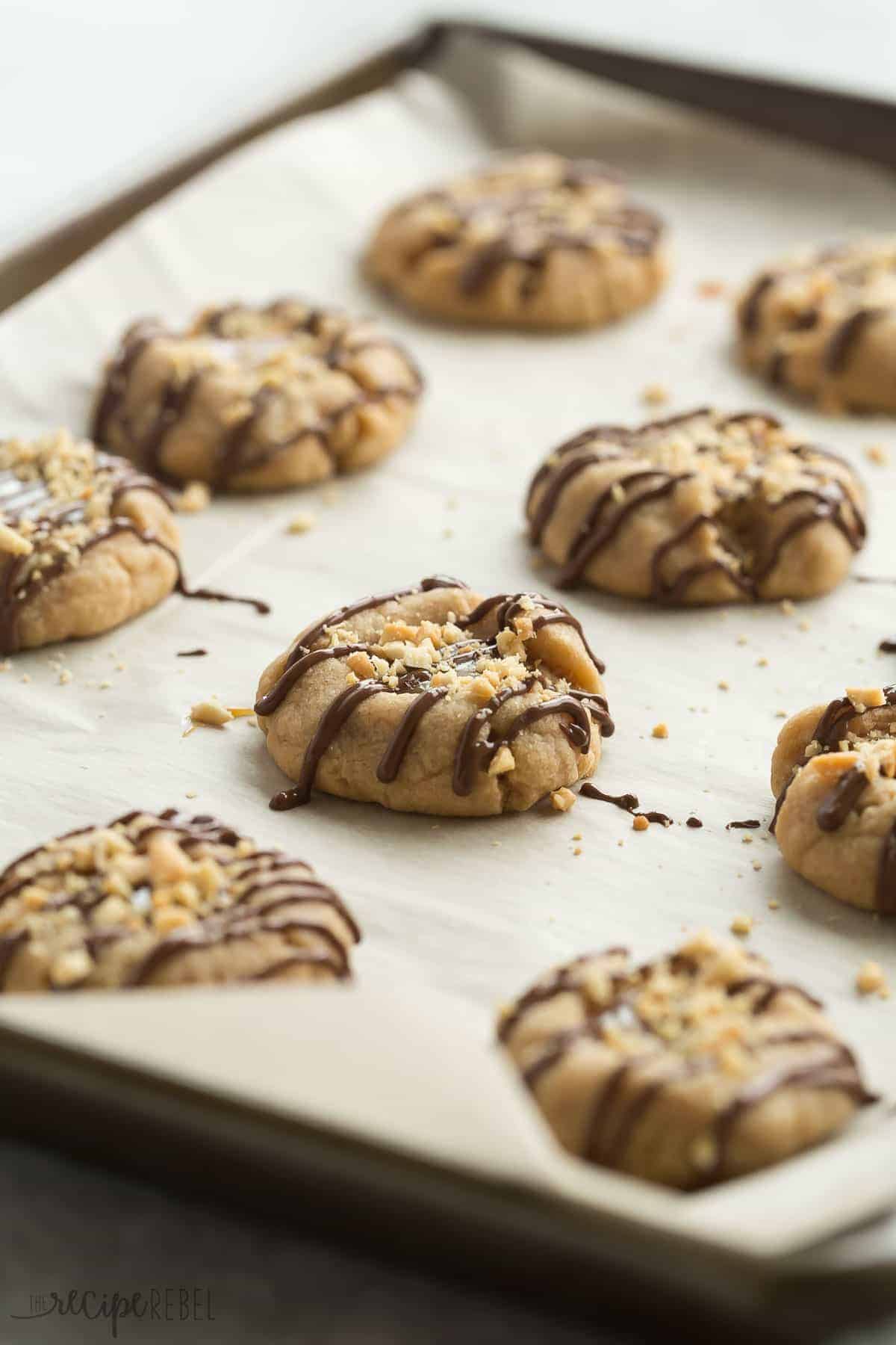 peanut butter turtle thumbprint cookies on a parchment lined sheet pan with chocolate drizzle