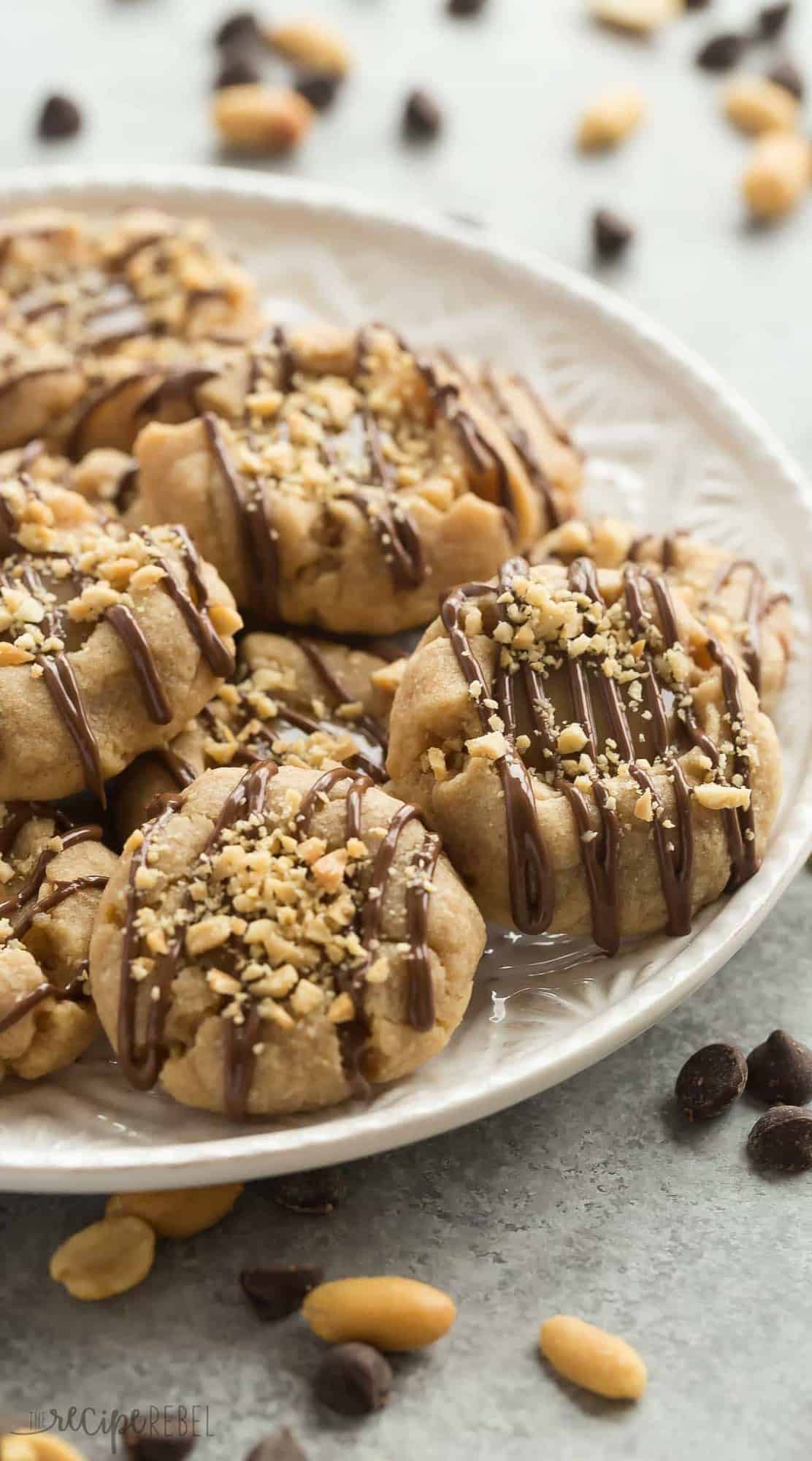 peanut butter thumbprint cookies on a white plate with chocolate drizzle and chopped peanuts