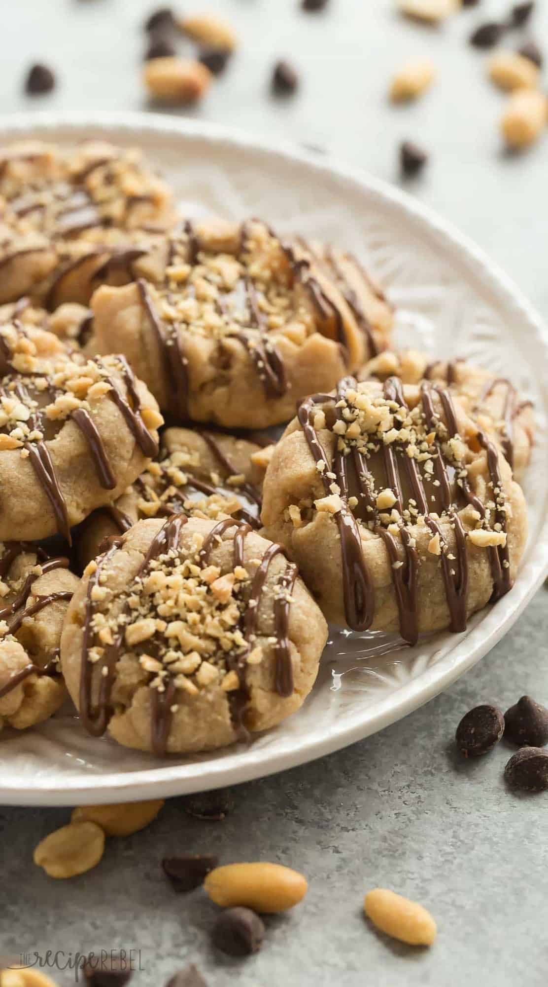 These Peanut Butter Turtle Thumbprint Cookies are a new twist on a classic Christmas cookie -- with a peanut butter cookie base, a simple caramel filling, a drizzle of chocolate and a sprinkling of peanuts, they're sure to be a new holiday tradition! peanut butter thumbprint cookies on a white plate with chocolate drizzle and chopped peanuts