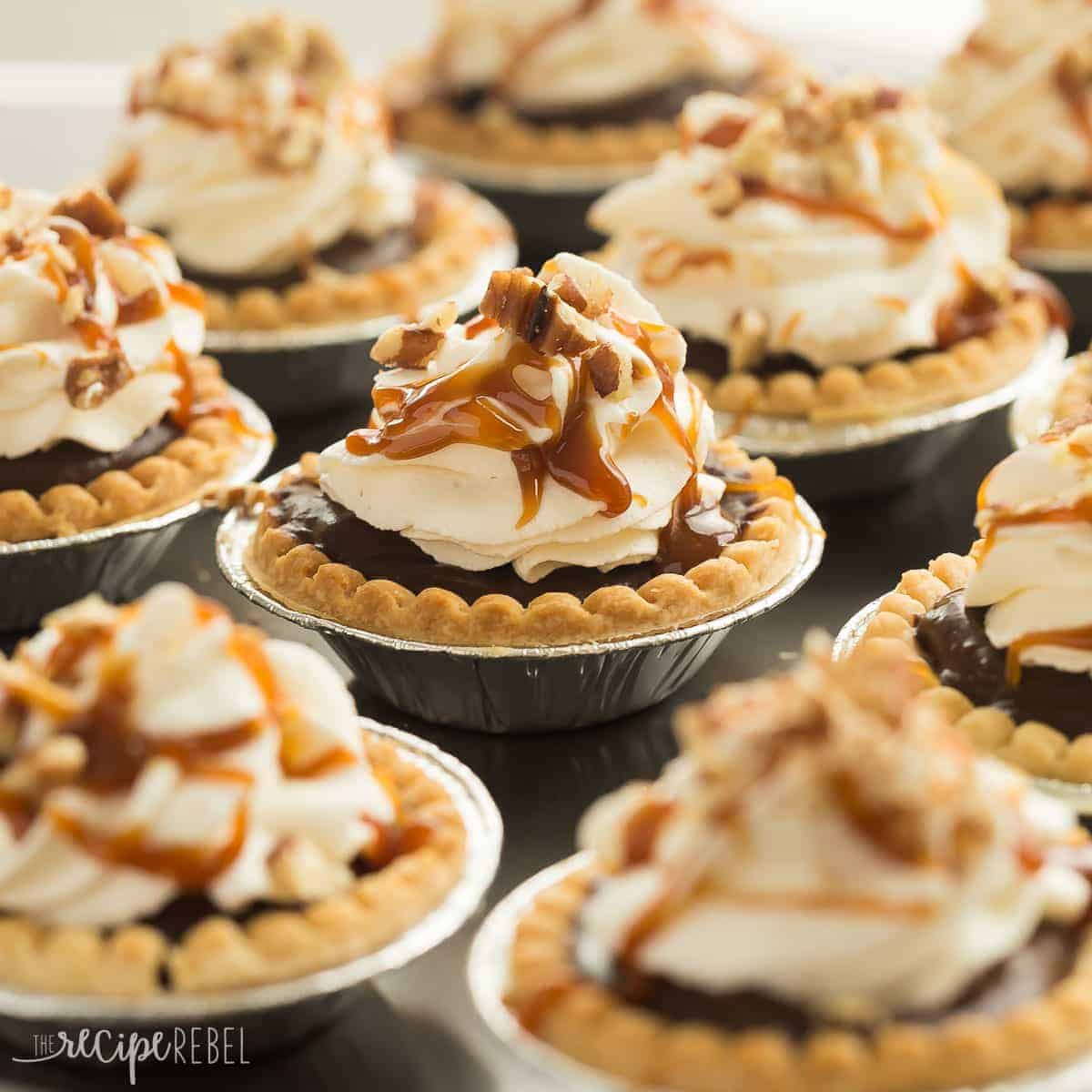 Turtle Pudding Pie Minis are the perfect bite-sized treat (made in tart shells!) for the holidays or any party -- they come together quickly and are loaded with pecans, caramel, chocolate pudding and topped with whipped cream! square image of many turtle pudding pies in tart shells with foil wrapper