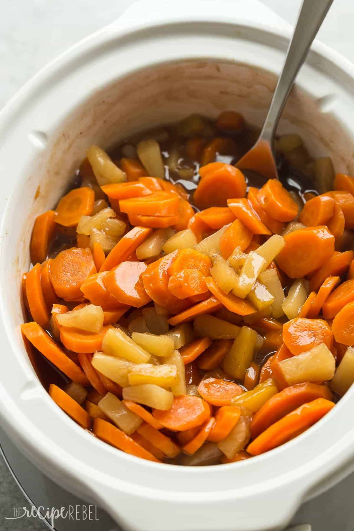 sliced carrots and pineapple in a slow cooker with metal spoon scooping