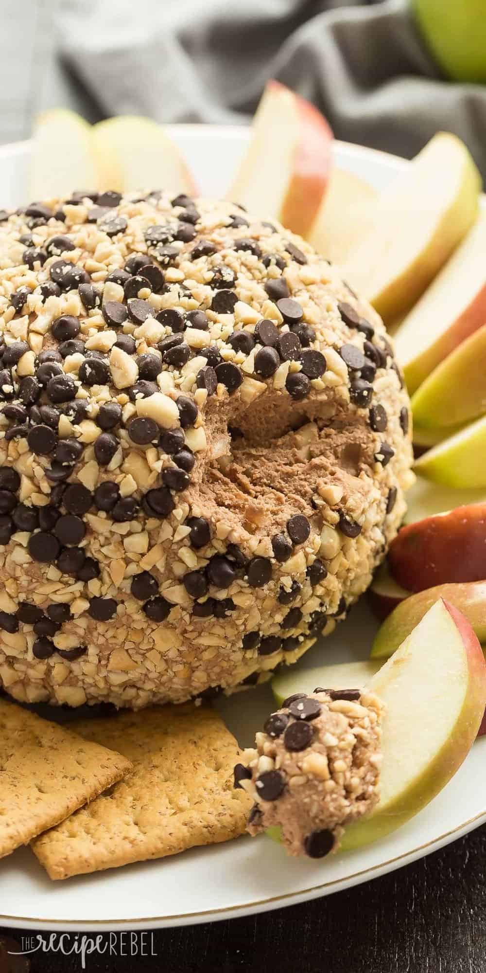 This Snickers Cheese Ball is an easy appetizer, snack or dessert -- filled with cream cheese, peanut butter, caramel and chocolate it is the BEST with apple slices! Perfect for potlucks. snickers cheese ball with apple dipped in cheese ball on plate