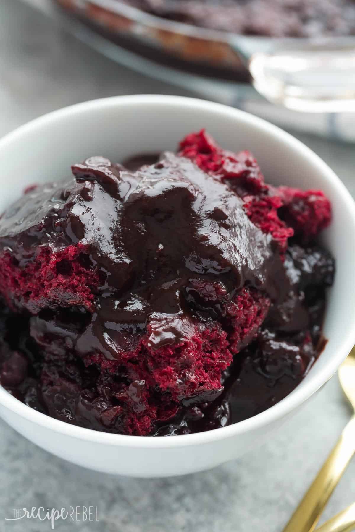 This RED VELVET Pudding Cake is so easy with only 10 minutes prep -- it has a chocolate cake base and it makes its own hot fudge sauce in the oven as it bakes and I highly recommend slathering it with a scoop of ice cream! Red velvet pudding cake in white bowl with pan in the background.
