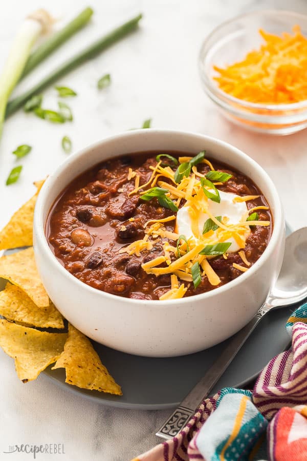 slow cooker chili in bowl close up with cheese green onions and chips in background