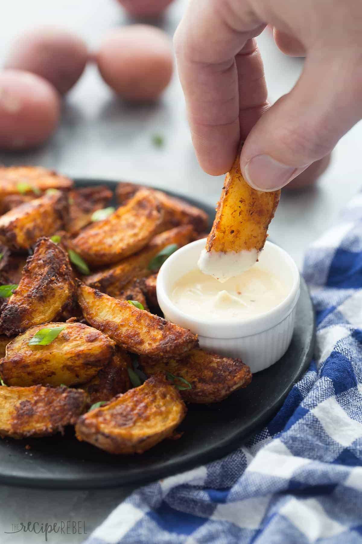 hand dipping chipotle parmesan potato wedge into small bowl of creamy sauce