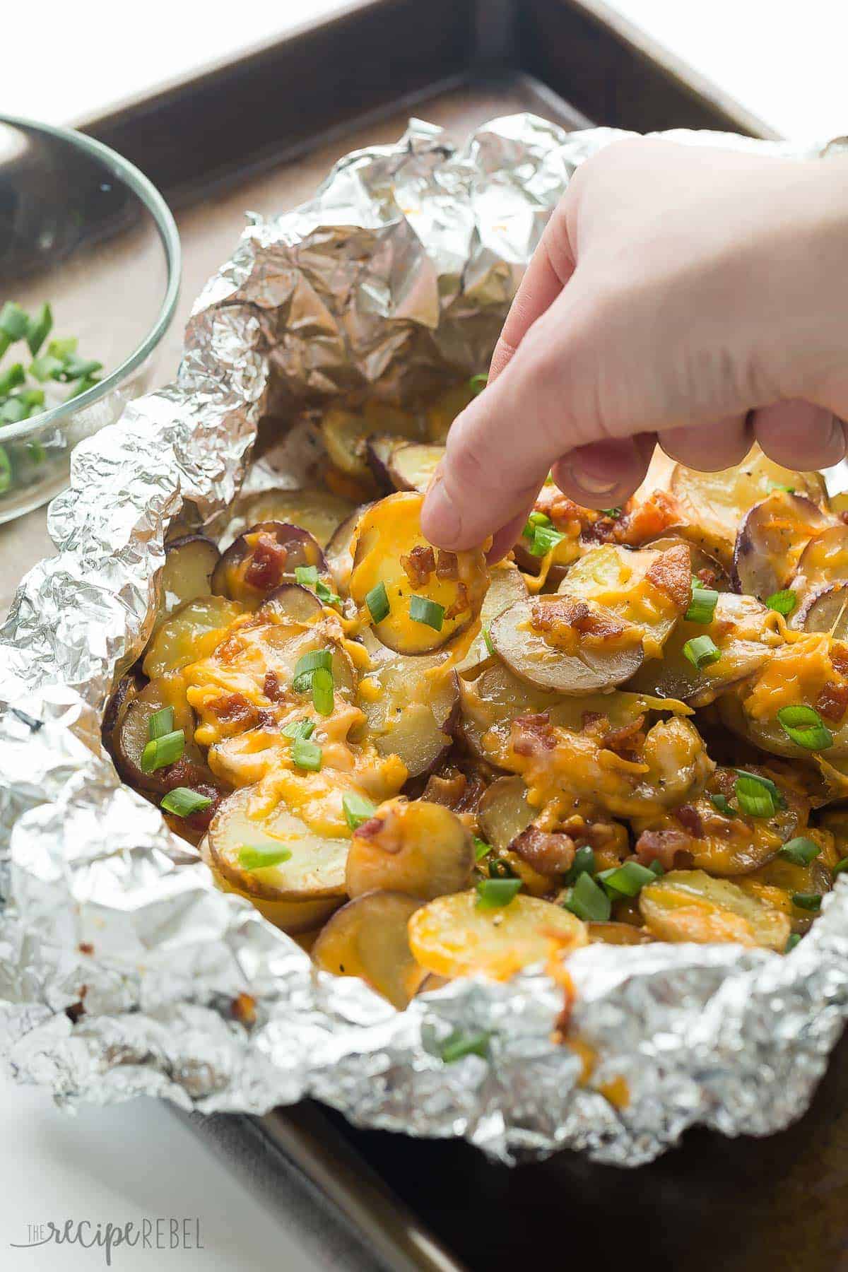 cheesy grilled potatoes in foil packet with bacon and green onions and hand pulling out potato