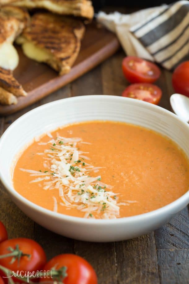 easy roasted tomato soup in white bowl with a sprinkle of shredded parmesan and grilled cheese in the background