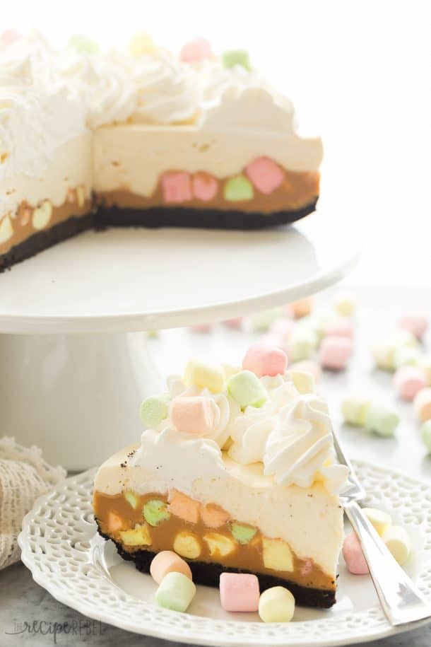 no bake peanut butter marshmallow square cheesecake slice on white plate with peanut butter marshmallow layer on the bottom and cheesecake layer on the top topped with whipped cream and colored marshmallows