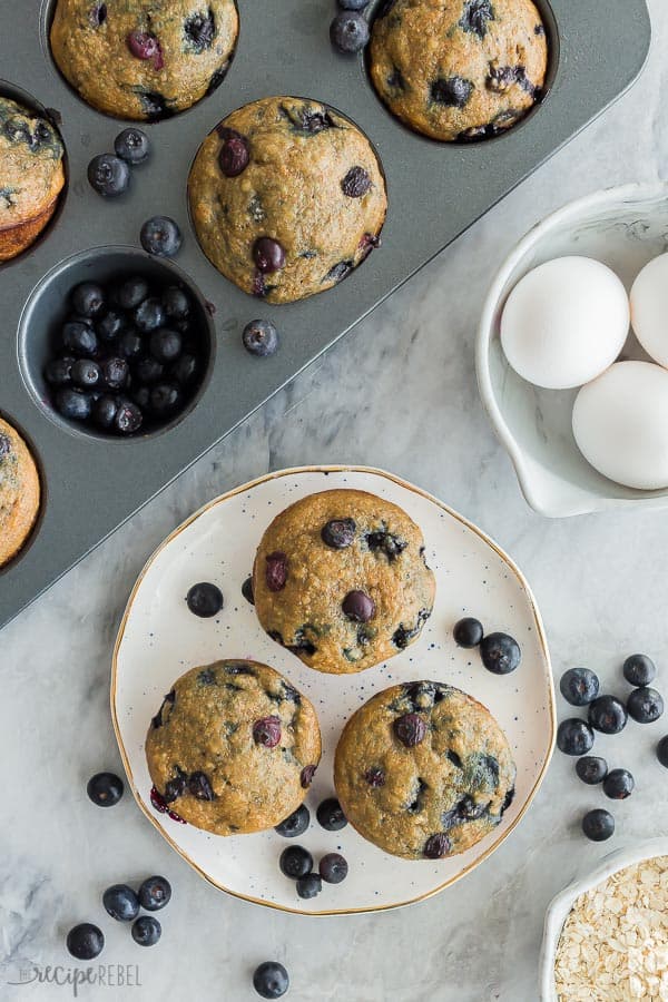 3 blueberry oatmeal muffins overhead on plate with fresh blueberries around the plate and fresh eggs in a bowl