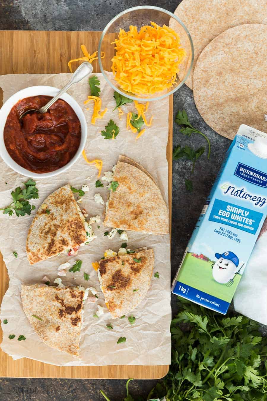 overhead image of breakfast quesadillas on wood cutting board with carton of egg whites on side