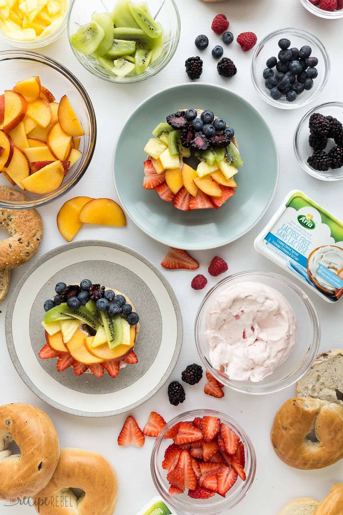 overhead image of rainbow fruit pizza bagels on plates with strawberry cream cheese and fruit all around