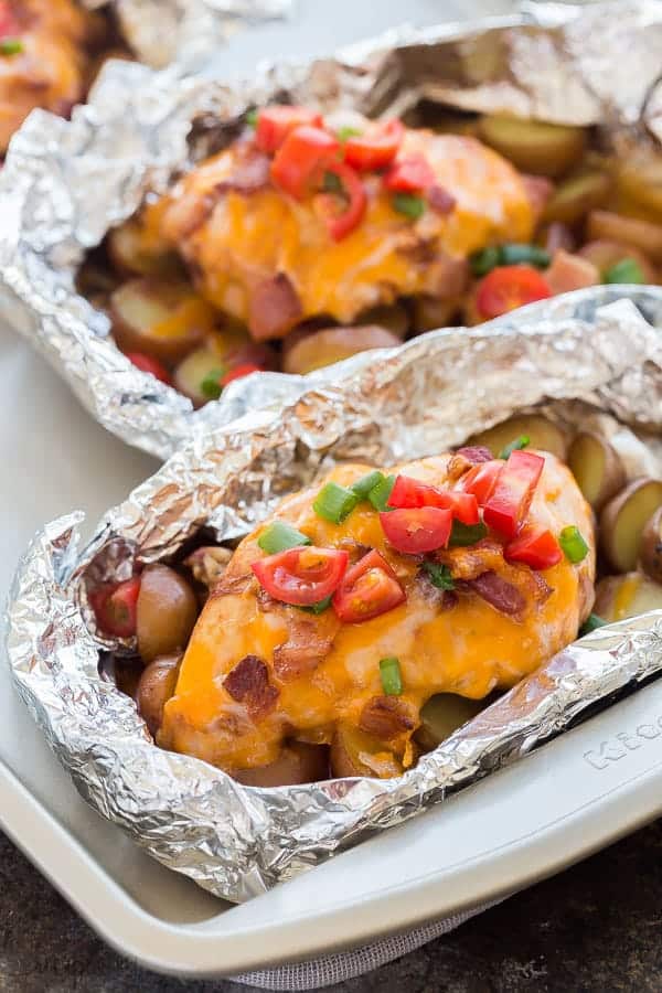 These Easy Monterey Chicken Foil Packets are the perfect easy dinner recipe for summer! You can make them in the oven or on the grill -- an easy camping meal! Sliced potatoes and vegetables topped with chicken breasts, barbecue sauce, cheese and bacon -- how can you go wrong?? #foilpack #foilpacket #chicken #dinner #recipe monterey chicken foil packs topped with cheddar cheese fresh tomatoes bacon and green onions on a sheet pan