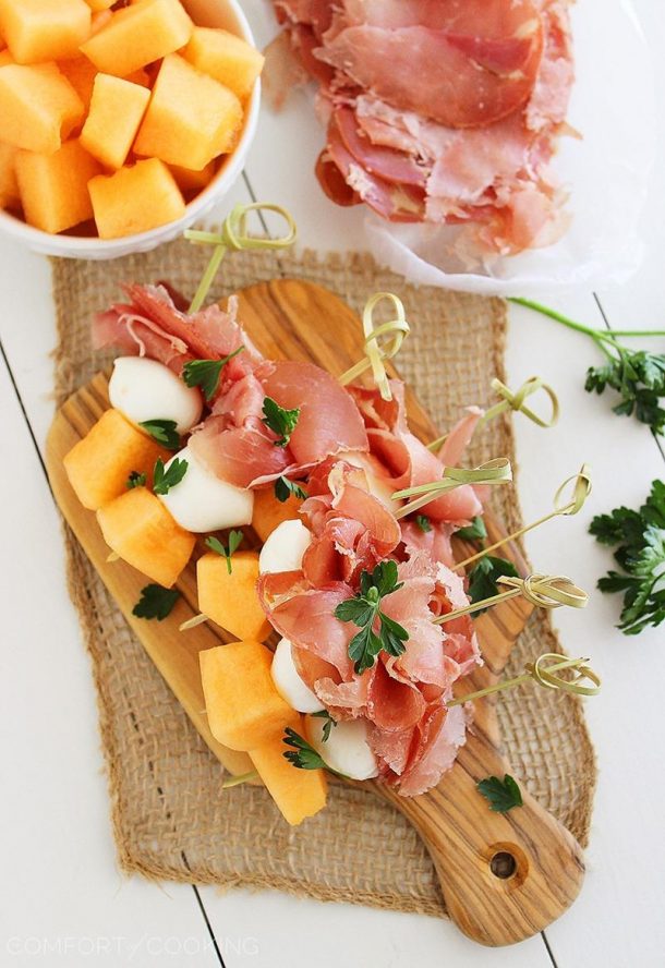 melon prosciutto and mozzarella skewers on gold skewers on top of wooden cutting board with melon chunks on the side