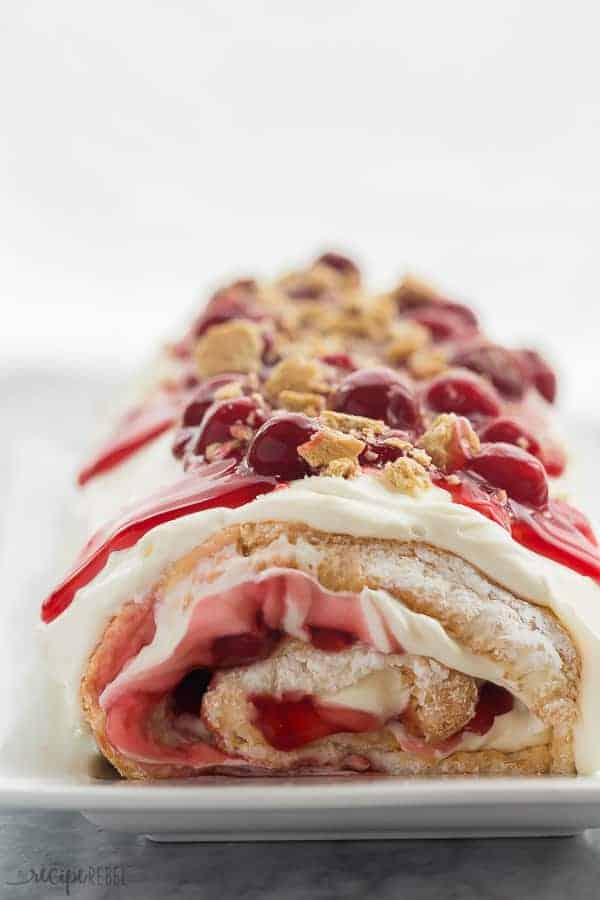 This Cherry Cheesecake Angel Food Cake Roll is the best kind of roll cake! It's made with light and fluffy angel food cake, and filled with a sweetened cream cheese whipped cream and cherry pie filling. It's the ULTIMATE summer dessert! Switch up the fruit to suit your tastes. Includes step by step RECIPE VIDEO #cherry #cherries #cheesecake #creamcheese #dessert #cake #angelfoodcake #recipe #recipes cherry cheesecake roll whole on a white plate with cherry pie filling and crushed graham crackers on top