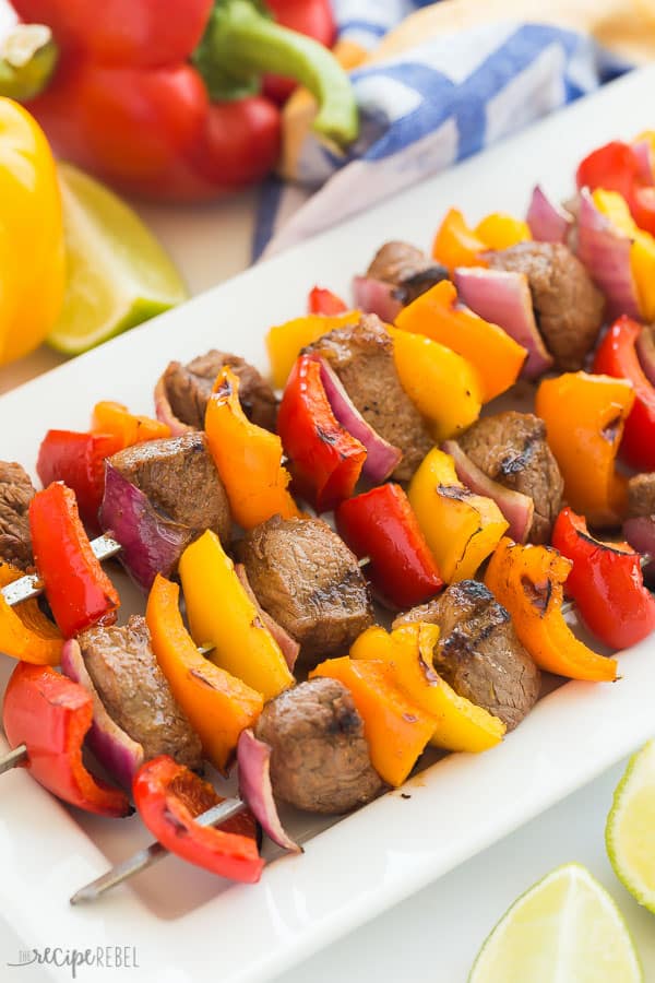 steak kabobs with peppers on a white plate with lime wedges and peppers in the background