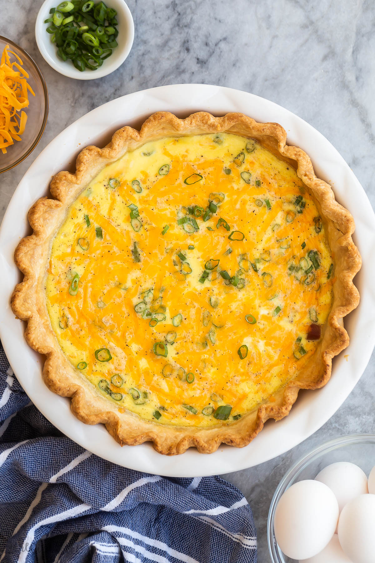 This Easy Ham and Cheese Quiche Recipe is made up of a rich, creamy, and cheesy egg custard that's baked into a buttery pie crust. It's packed with ham, cheddar cheese, and green onions. Just 15 minutes to prep! #breakfast #dinner #recipes | easy recipes | breakfast ideas | healthy recipes | easter brunch | holiday breakfast | eggs overhead image of whole ham quiche on marble background