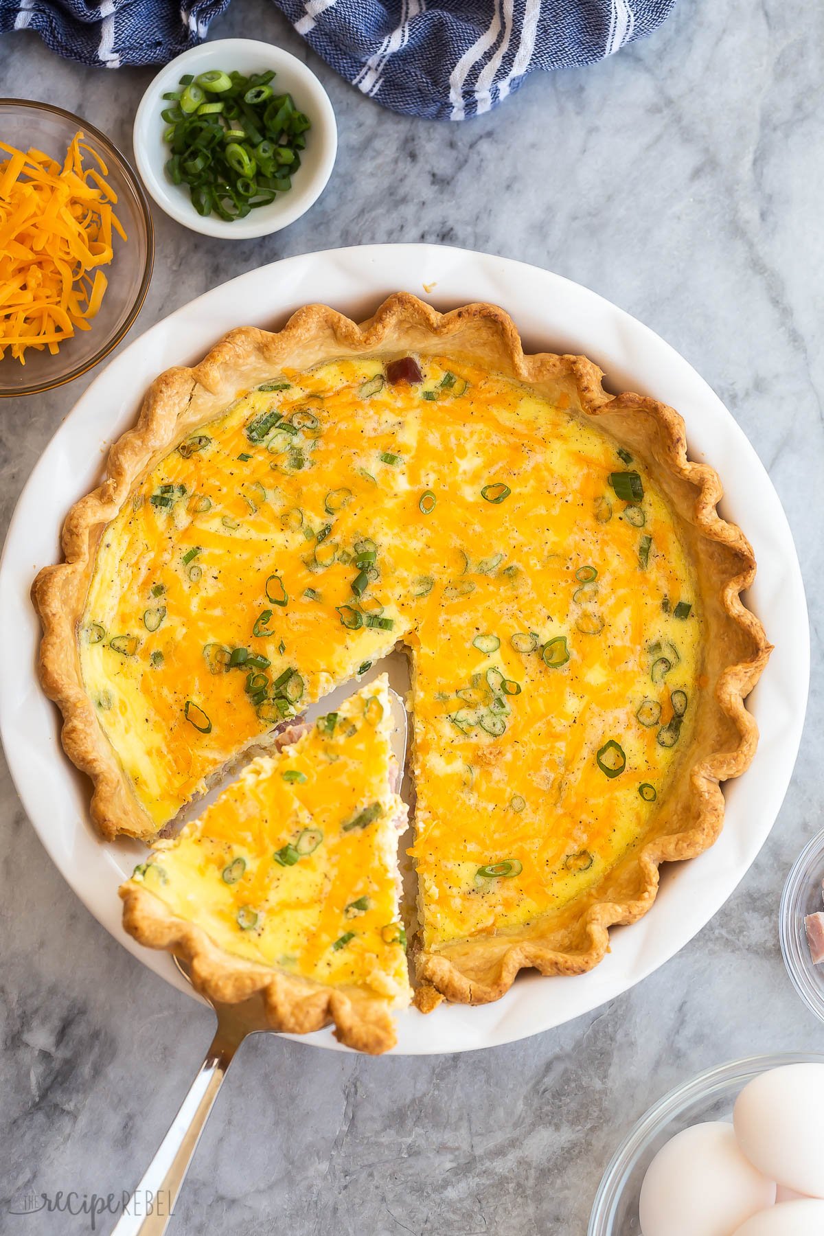 This Easy Ham and Cheese Quiche Recipe is made up of a rich, creamy, and cheesy egg custard that's baked into a buttery pie crust. It's packed with ham, cheddar cheese, and green onions. Just 15 minutes to prep! #breakfast #dinner #recipes | easy recipes | breakfast ideas | healthy recipes | easter brunch | holiday breakfast | eggs piece of ham quiche being lifted out of whole from above