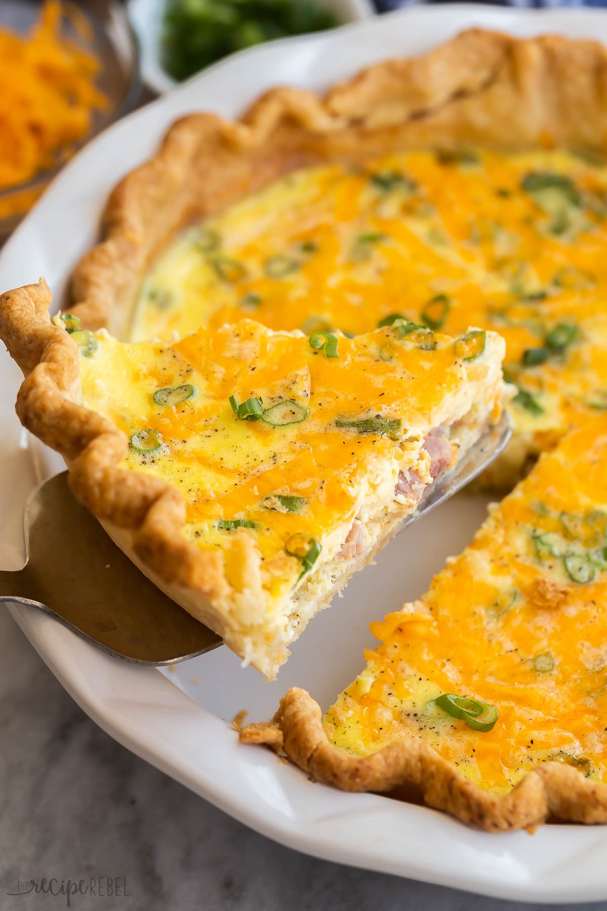 This Easy Ham and Cheese Quiche Recipe is made up of a rich, creamy, and cheesy egg custard that's baked into a buttery pie crust. It's packed with ham, cheddar cheese, and green onions. Just 15 minutes to prep! #breakfast #dinner #recipes | easy recipes | breakfast ideas | healthy recipes | easter brunch | holiday breakfast | eggs piece of ham quiche being lifted out of pie plate