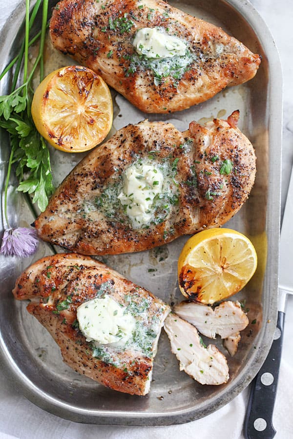 grilled chicken breasts overhead on metal pan with chive butter on top and lemon halves on the side