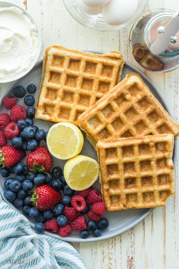ricotta yeast waffles overhead on a large grey plate with lemon halves and fresh berries on a rustic white background