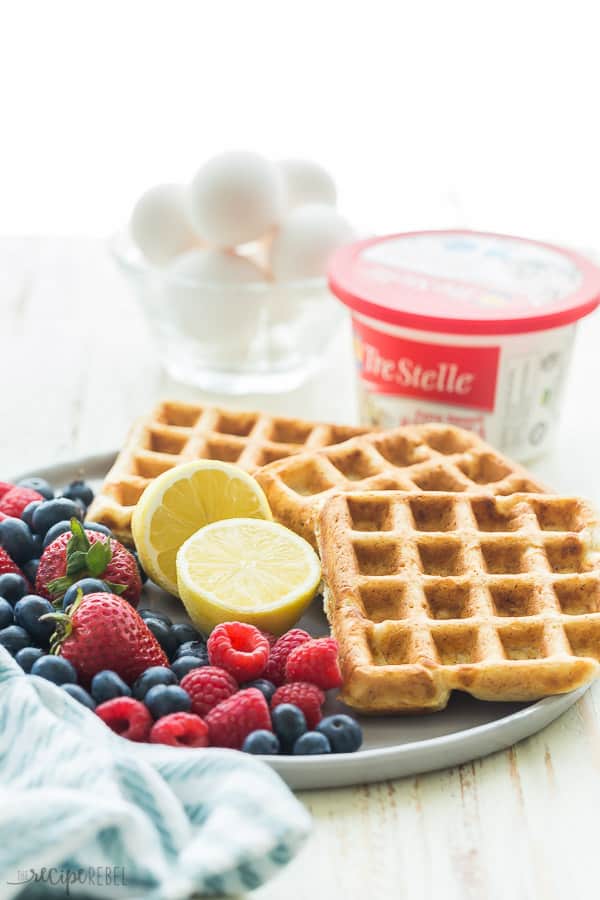 lemon ricotta waffles on a plate with lemon halves fresh berries and a tub of ricotta cheese in the background