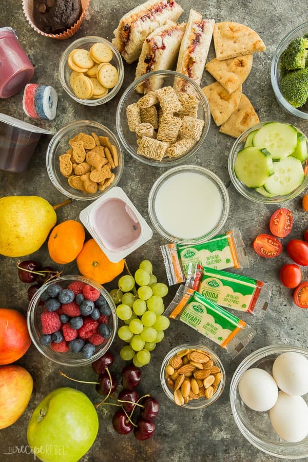 school lunch ideas overhead with dry cereal lots of different fruits crackers cheese bars nuts whole eggs raw vegetables and sandwiches