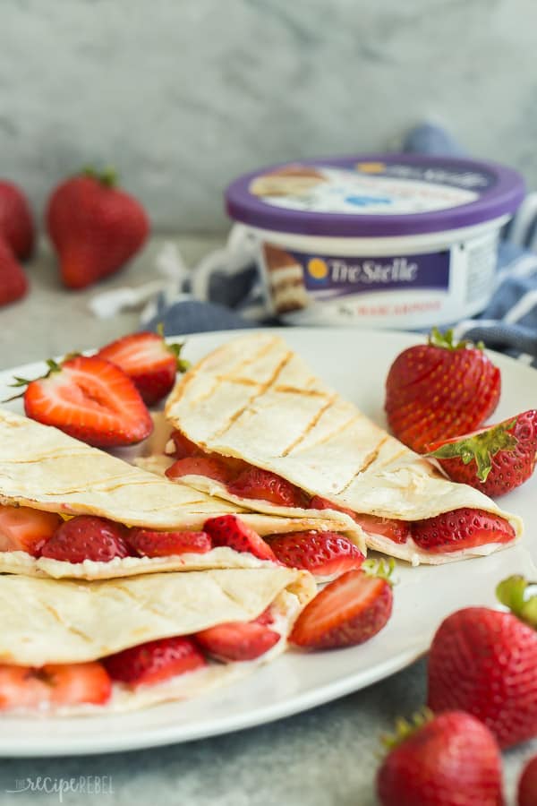 These Strawberry Cheesecake Quesadillas are a simple grilled dessert recipe that is easy to prepare and tastes incredible! Switch up the fruit and flavours to make your own variations, or take them to the stovetop to enjoy all year long. #cheesecake #strawberry #strawberries #quesadilla #dessert #recipe strawberry quesadillas on white plate with tre stelle mascarpone tub in the background and lots of fresh strawberries around