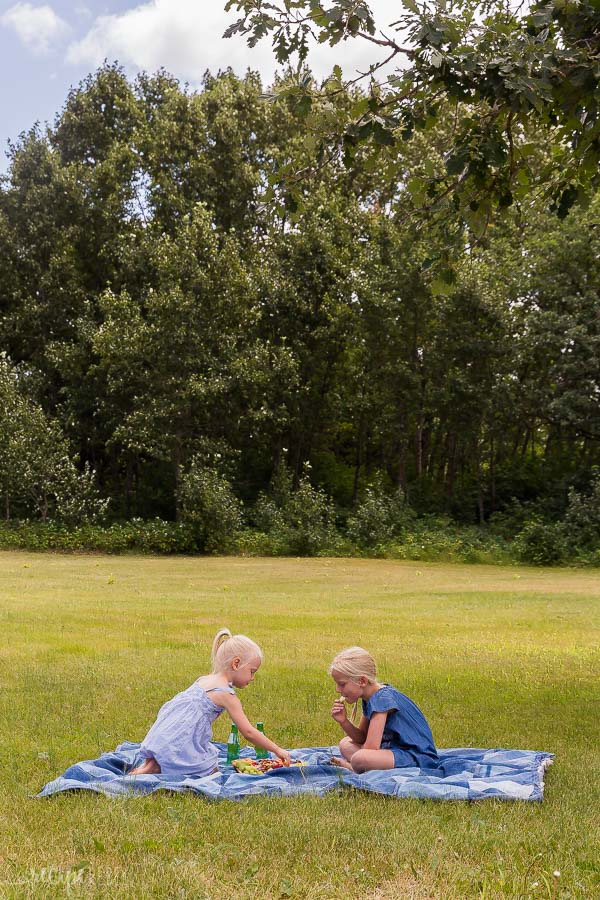 two young girls having a picnic on a denim picnic blanket with trees in the background