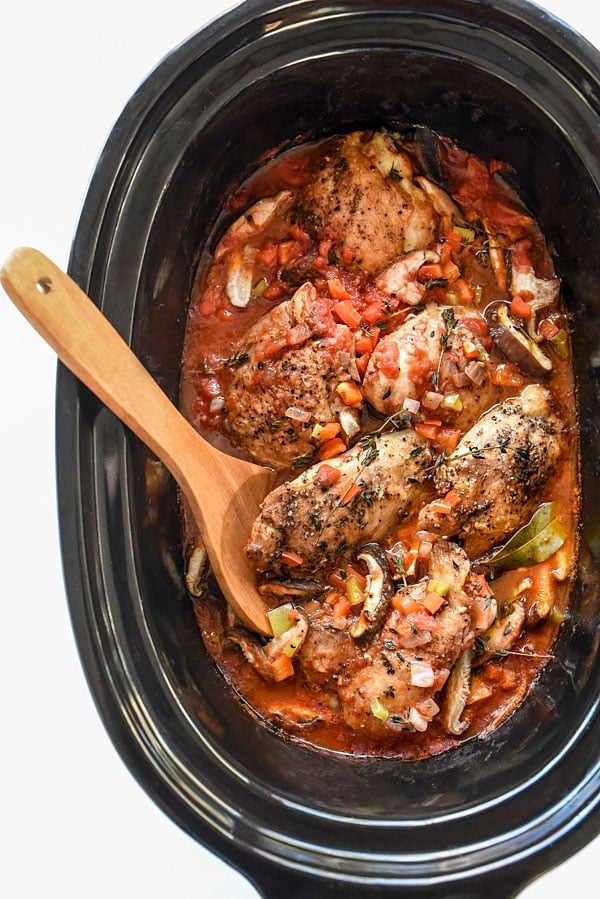 slow cooker chicken cacciatore overhead in a large black crockpot with a wooden spoon stuck into the crockpot