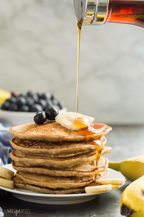 These Easy Banana Oatmeal Pancakes are made with soaked oats and mashed bananas, which gives them incredible moisture and an extra boost of fiber! They're an easy, healthy pancake recipe that is freezer friendly and perfect for school mornings! Includes step by step recipe video. #pancakes #breakfast #banana #blueberry #recipe #healthy banana oat pancakes with syrup being drizzled onto stack with fresh blueberries and banana slices in the background