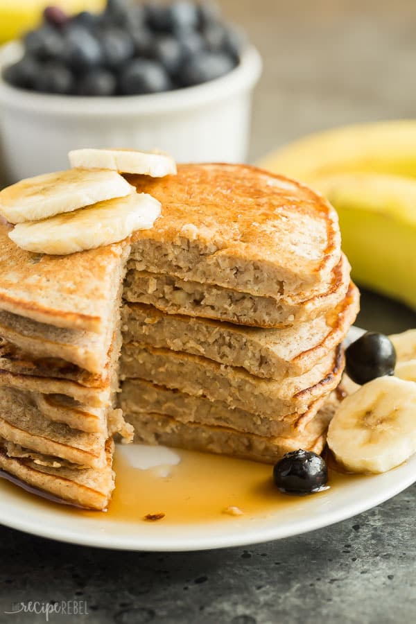 These Easy Banana Oatmeal Pancakes are made with soaked oats and mashed bananas, which gives them incredible moisture and an extra boost of fiber! They're an easy, healthy pancake recipe that is freezer friendly and perfect for school mornings! Includes step by step recipe video. #pancakes #breakfast #banana #blueberry #recipe #healthy banana oatmeal pancakes stack on a plate with one wedge cut out of stack and syrup on plate