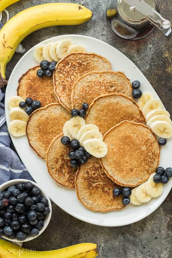 banana oatmeal pancakes on a platter with fresh blueberries and banana slices all around on a dark grey background