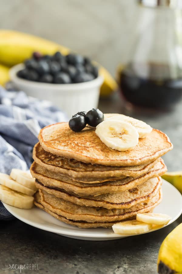 These Easy Banana Oatmeal Pancakes are made with soaked oats and mashed bananas, which gives them incredible moisture and an extra boost of fiber! They're an easy, healthy pancake recipe that is freezer friendly and perfect for school mornings! Includes step by step recipe video. #pancakes #breakfast #banana #blueberry #recipe #healthy banana pancakes stack on white plate with fresh blueberries and banana slices on top