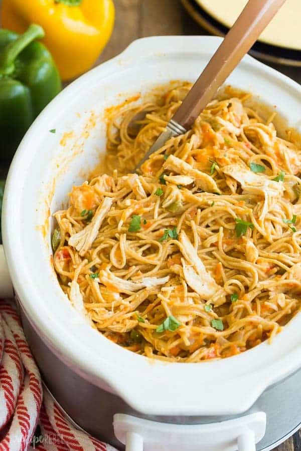 crockpot chicken spaghetti in white slow cooker with metal spoon scooping pasta and whole peppers in the background