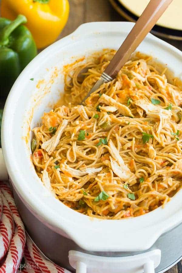 This Cheesy Crockpot Chicken Spaghetti is an easy crockpot meal for busy weeknights! It's made healthier with no canned soups, lean chicken breast and added veggies, and it cooks completely in the slow cooker for less clean up! Includes step by step recipe video. #crockpot #slowcooker #spaghetti #pasta #chicken #chickenrecipe crockpot chicken spaghetti in white slow cooker with metal spoon scooping pasta and whole peppers in the background
