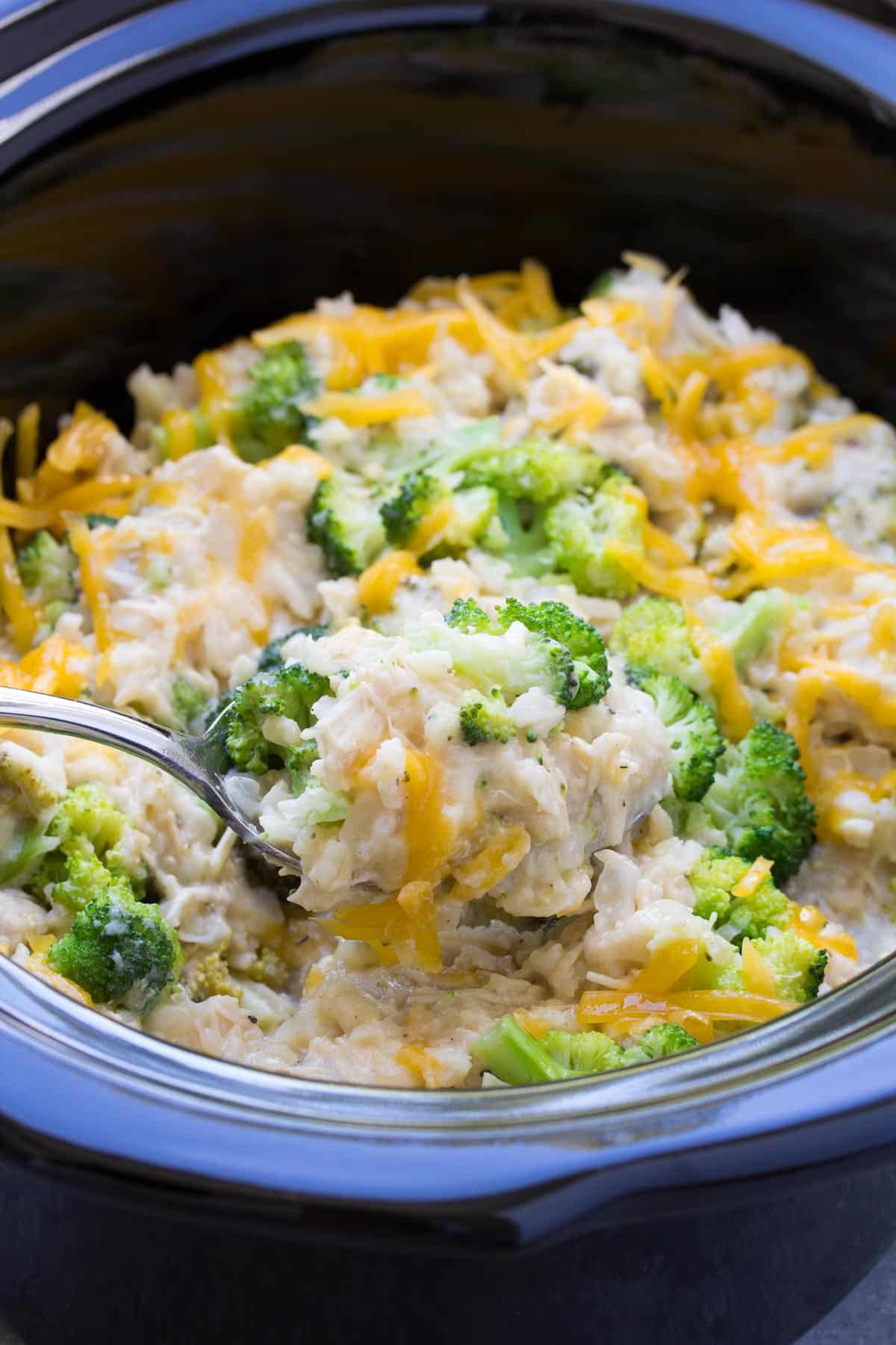 slow cooker chicken broccoli rice casserole in black crockpot with a metal spoon scooping some up