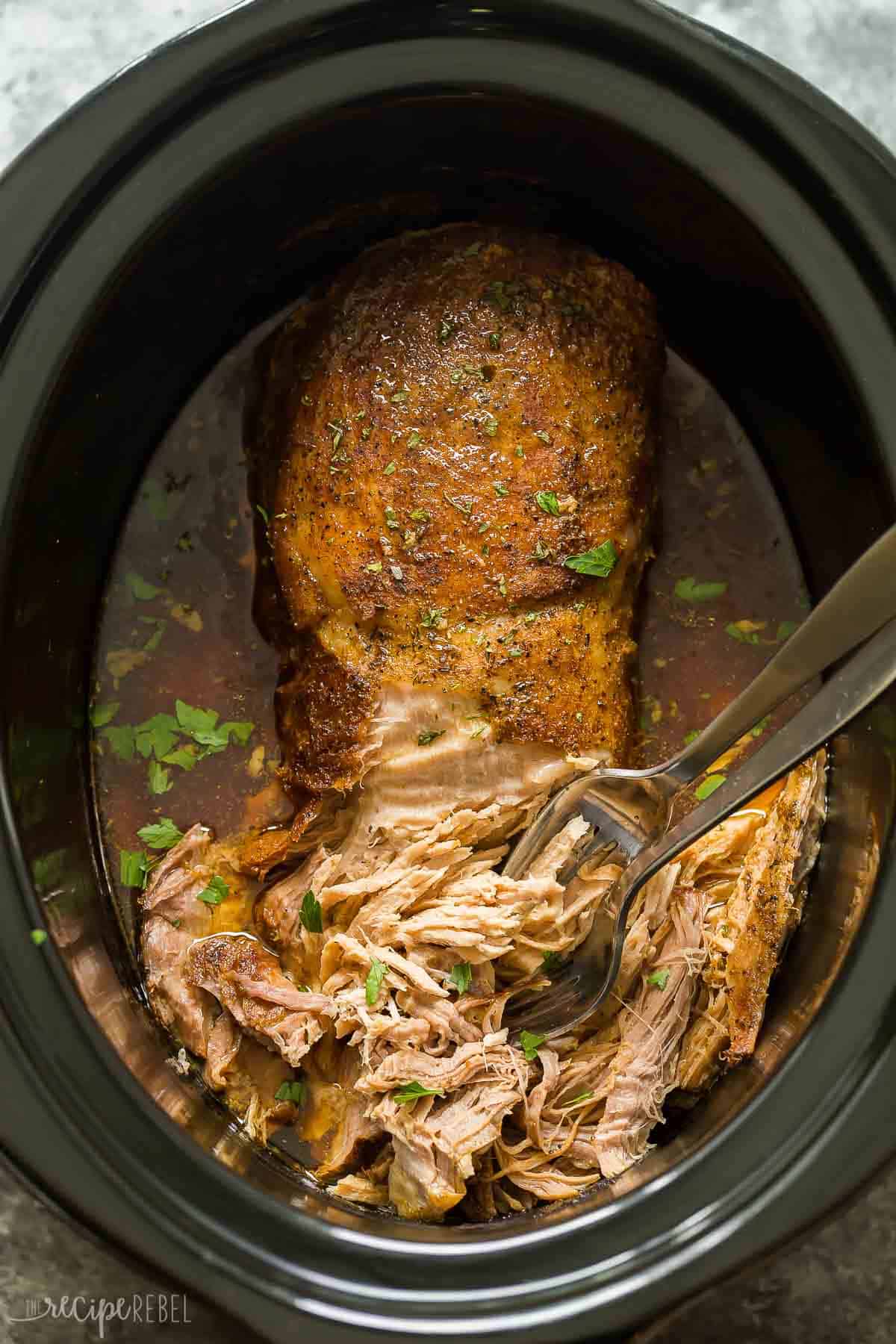 This easy Slow Cooker Pork Loin recipe includes a flavorful garlic herb rub, and is slow cooker in apple juice for extra flavor! It is fall apart tender with minimal effort! #slowcooker #crockpot #recipe #dinner #pork overhead image of pork loin in black crockpot with two forks to shred.