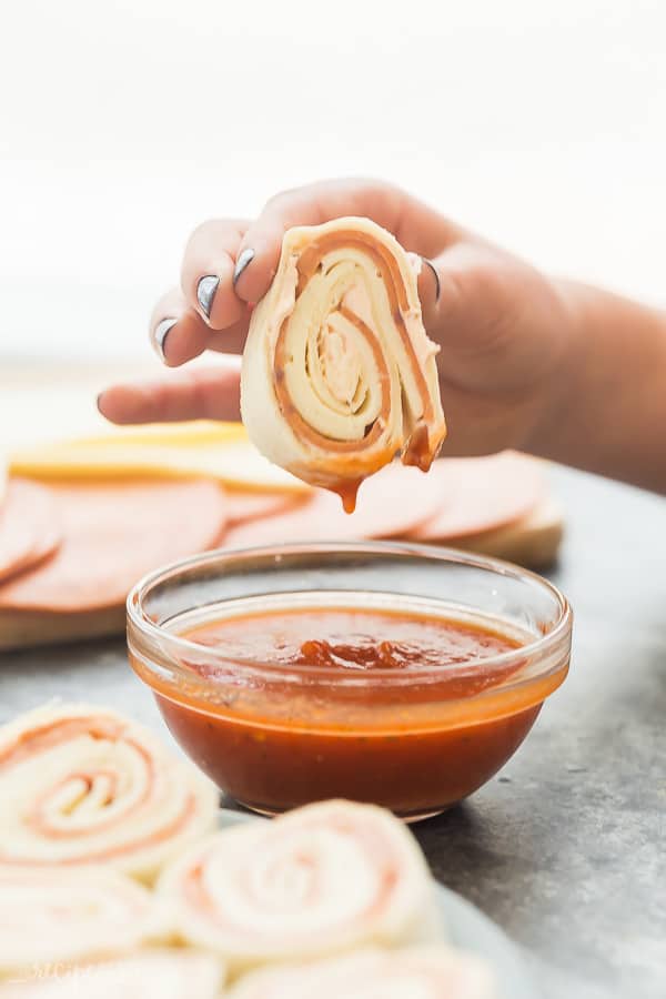 These Tortilla Roll Ups are made PIZZA style, with a tomato sauce and cream cheese spread, sliced pepperoni and most importantly -- cheese! An easy make ahead lunch idea. Includes step by step recipe video. #rollups #snack #appetizer #lunch #pizza #recipe tortilla roll ups dipped in small glass bowl of pizza sauce