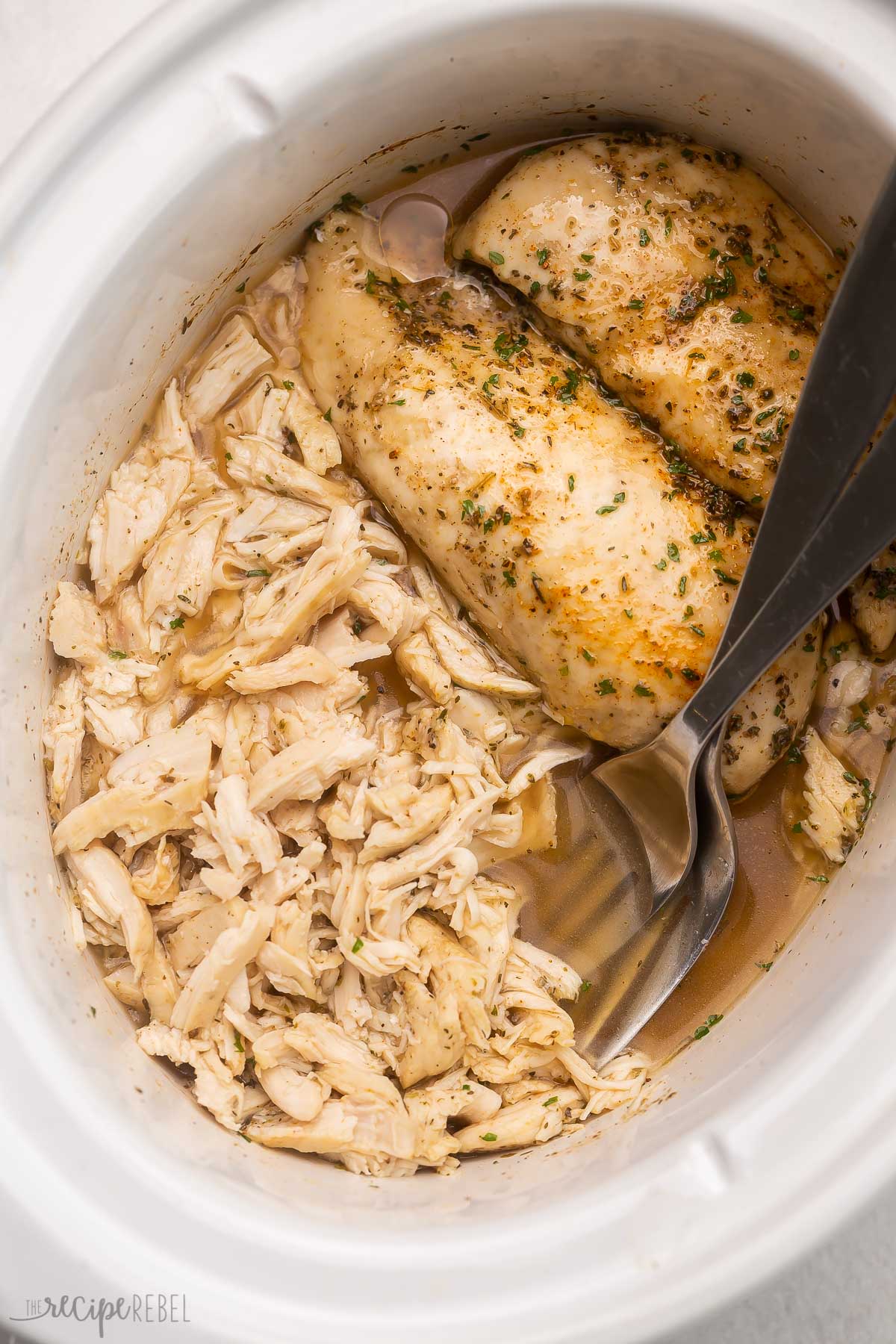 two whole chicken breasts and two shredded chicken breasts with forks in slow cooker.
