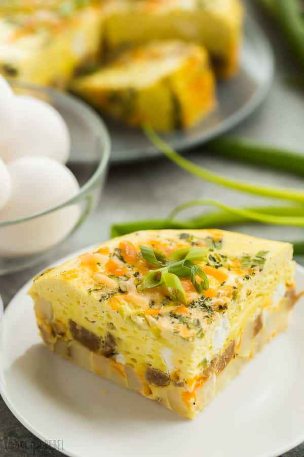 slice of sausage egg casserole on white plate with whole eggs and green onions in the background