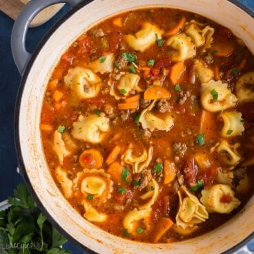 square image of sausage tortellini soup in grey dutch oven on blue background.
