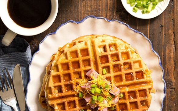 Top view of a ham and cheese waffle on a plate, next to coffee mugs.