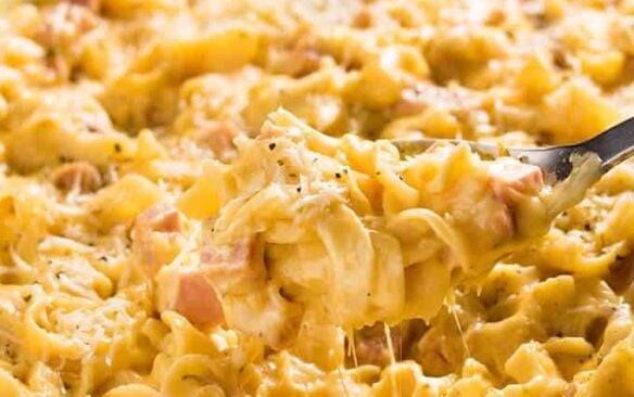 A scoop of cheesy ham and noodle casserole is lifted from a baking dish.