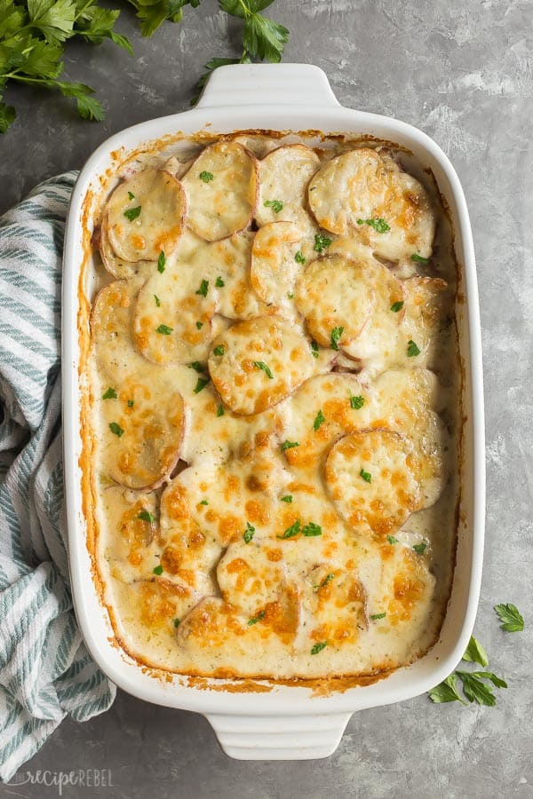 These Cheesy Scalloped Potatoes and Ham are an easy holiday dinner idea that's also a great recipe for leftover ham! The whole family is going to love this one! #potatoes #casserole #recipe #dinner #ham cheesy scalloped potatoes and ham in white dish on grey background with blue striped towel on the side