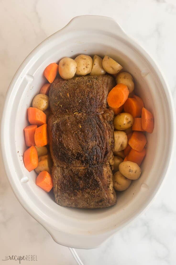 browned beef roast in white slow cooker with potatoes and carrots