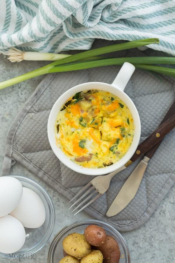 This Breakfast in a Mug recipe is so quick to whip up on a busy morning! You can cook it in the microwave or bake it in a baking dish. Easy and healthy! #breakfast #healthy #recipe #lunch #eggs easy breakfast casserole in a white mug overhead on grey towel with eggs and green onions in the background
