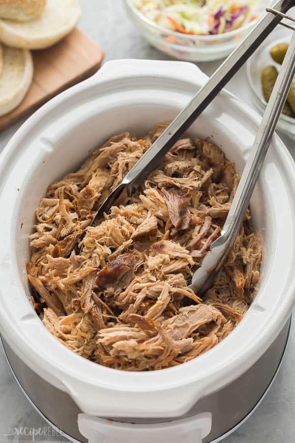 crock pot pulled pork in slow cooker with metal tongs stuck in and buns in the background