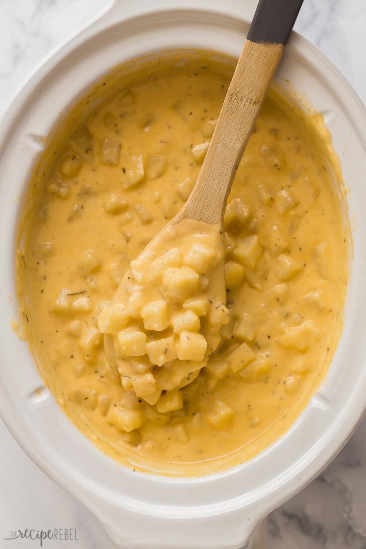 overhead image of wooden spoon lifting cheesy potatoes out of crockpot