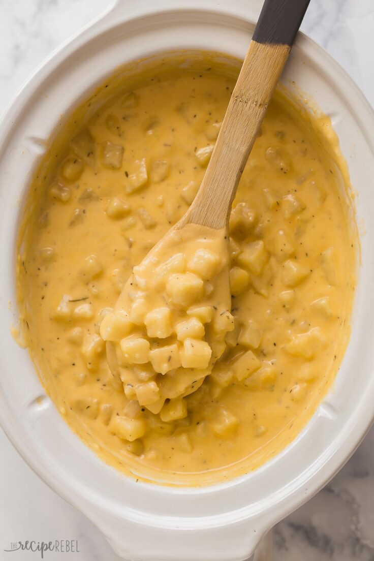 overhead image of wooden spoon lifting cheesy potatoes out of crockpot