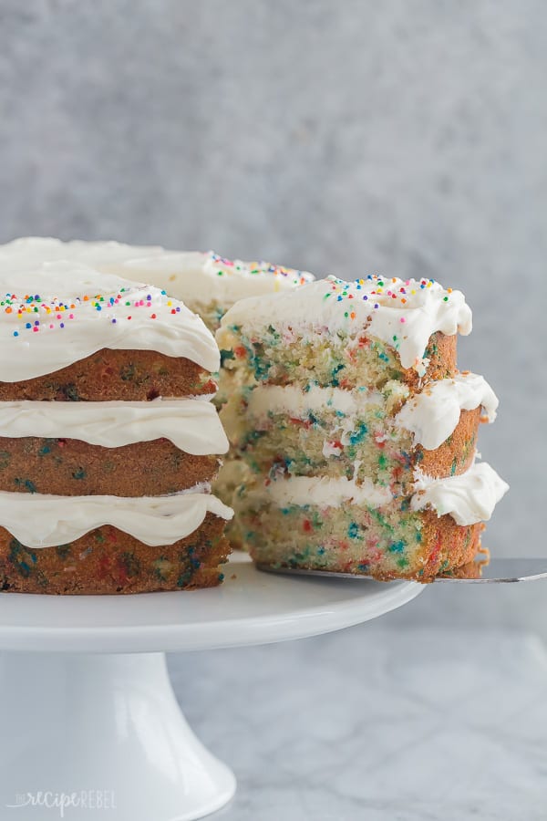 funfetti cake sitting on white cake plate with slice coming out on grey background